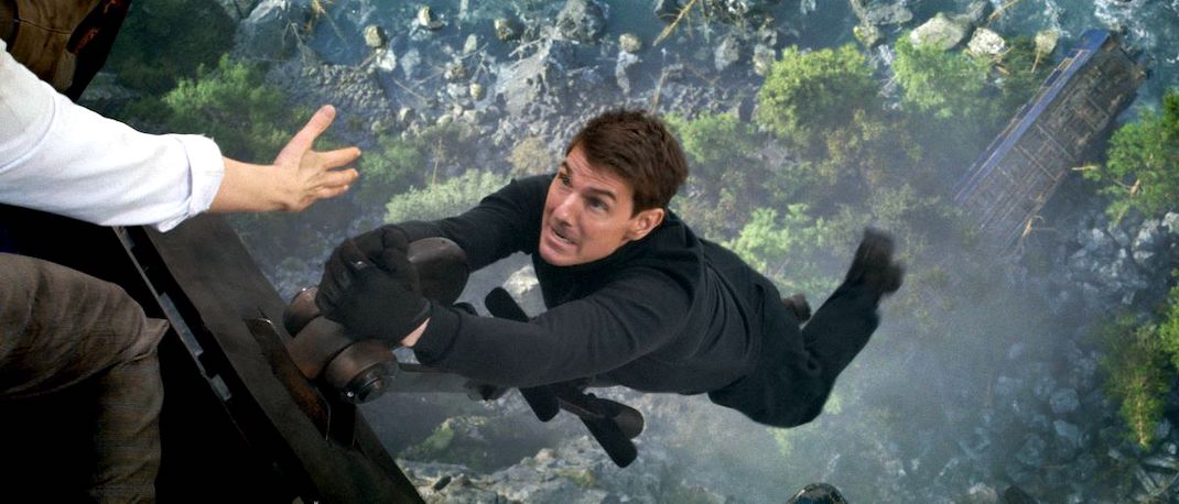 Tom Cruise hangs by his fingertips from a moving train, over a rocky chasm, while a hand reaches out to help him, in Tom Cruise in MISSION IMPOSSIBLE DEAD RECKONING PART ONE